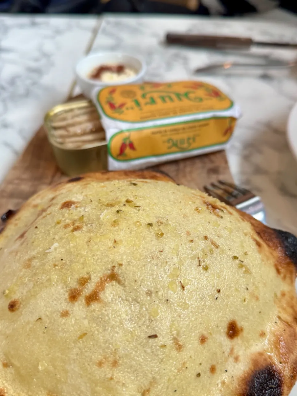 A puffed dome of pita bread on a wooden board at Kallos, glossy with olive oil and dusted with oregano, charred and blistered around the edge, with a Nuri spiced mackerel tin and a small dish of aioli behind it.