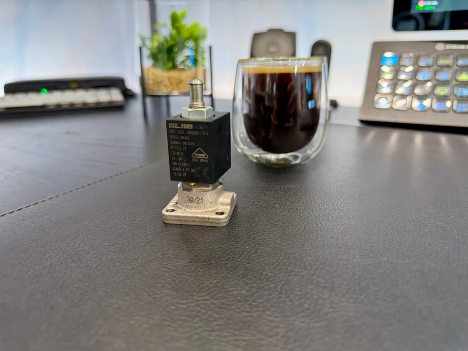 The coffee machine's solenoid valve lying on a wooden desk beside a cup of coffee.